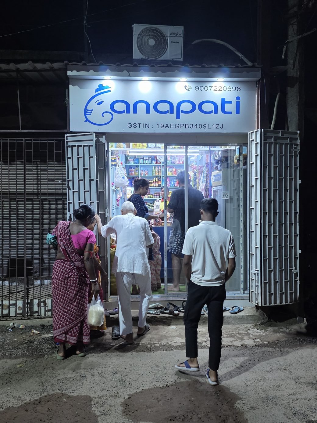 The Ganapati storefront at night — the lit signboard above open gates, with customers shopping inside.